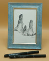 Framed black and white drawing of a natural landscape with two pens on a beige background