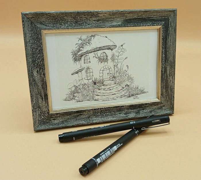 Framed black and white drawing of a fairy house with a mushroom shaped roof and two black markers on a beige background