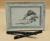 Framed black and white drawing of trees on rocky islands on a beige background with two pens below.