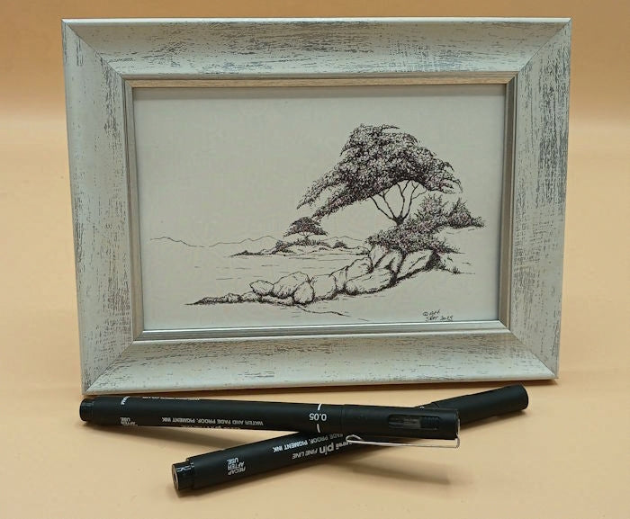 Framed black and white drawing of trees on rocky islands on a beige background with two pens below.