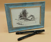Framed black and white drawing of a landscape featuring trees and rock outcrops with two pens on a beige background
