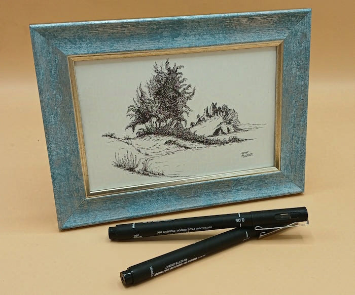 Framed black and white drawing of a landscape featuring trees and rock outcrops with two pens on a beige background