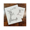 Two white paper prints with black floral letter 'E' designs on a wooden surface