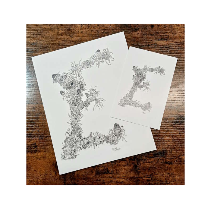 Two white paper prints with black floral letter 'E' designs on a wooden surface